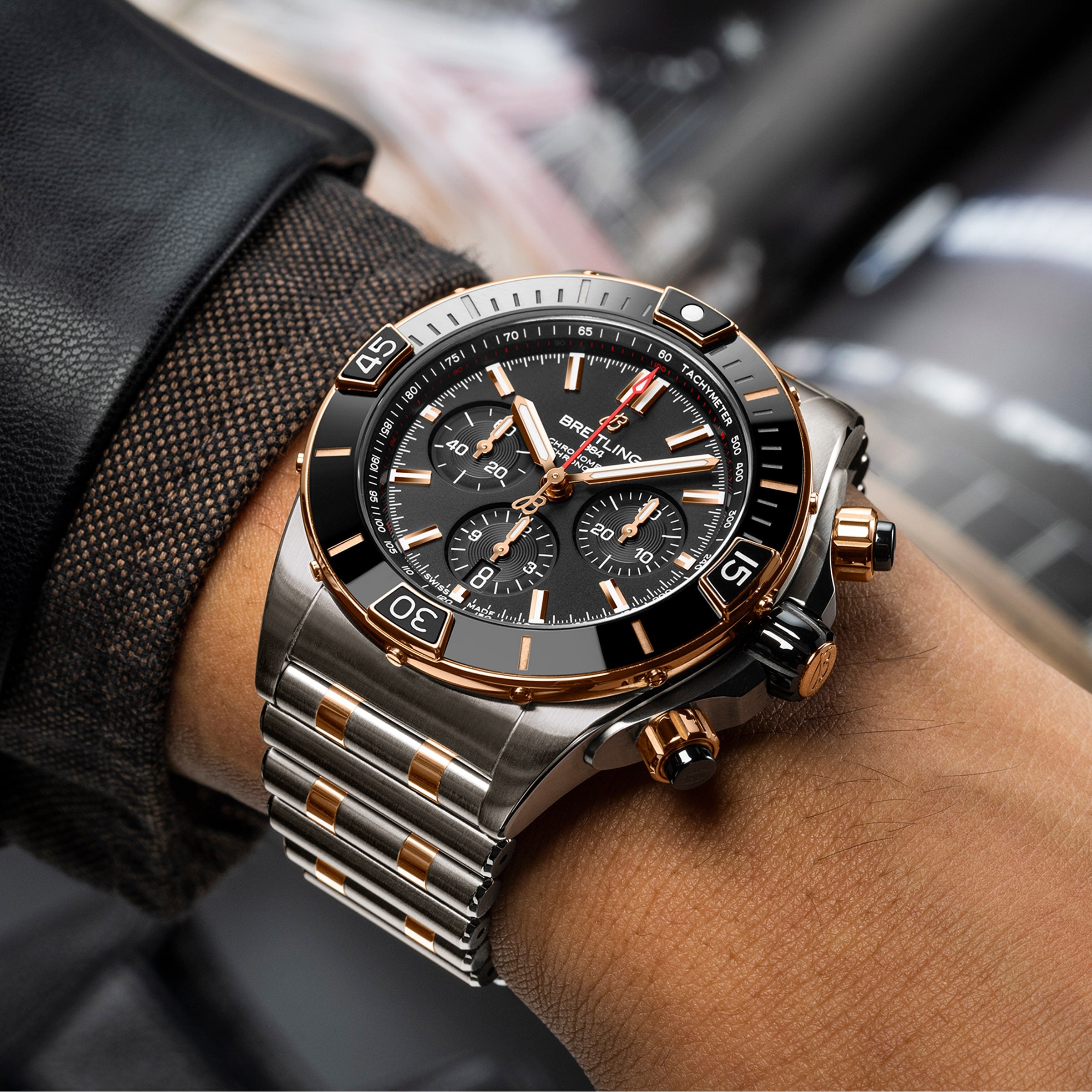 A close-up of a wrist wearing the Breitling Super Chronomat B01 44, a COSC-certified chronograph with a black dial, 18ct rose gold accents, and a two-tone metal bracelet.