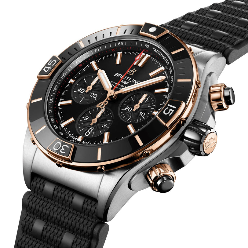 The Breitling Super Chronomat B01 44 is a luxury mechanical chronograph watch featuring a black dial, gold accents, three subdials, a black bezel, and a textured black strap.