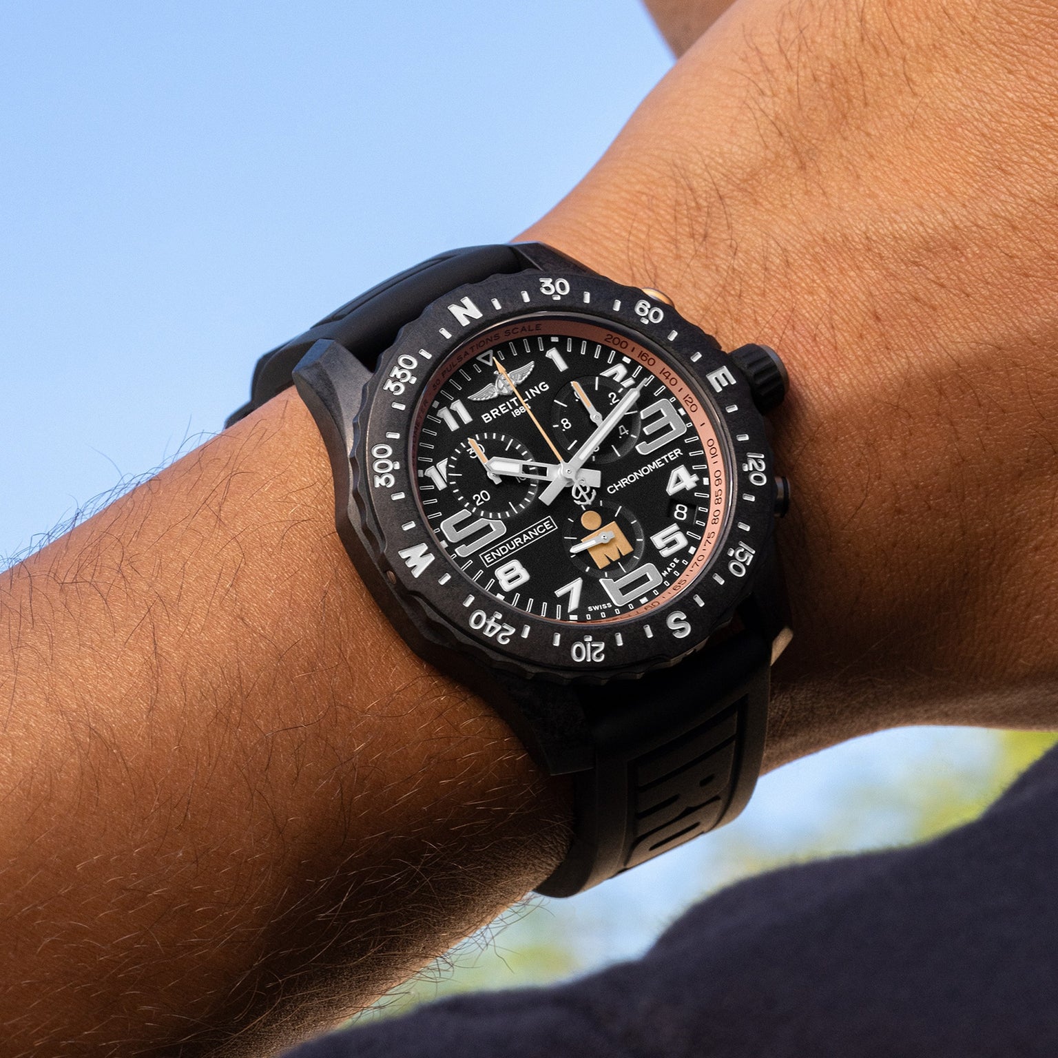 A close-up of a wrist wearing the black Breitling Endurance Pro 44 IRONMAN® Finisher watch with a black rubber strap, crafted in lightweight Breitlight®, set against a blue sky background.
