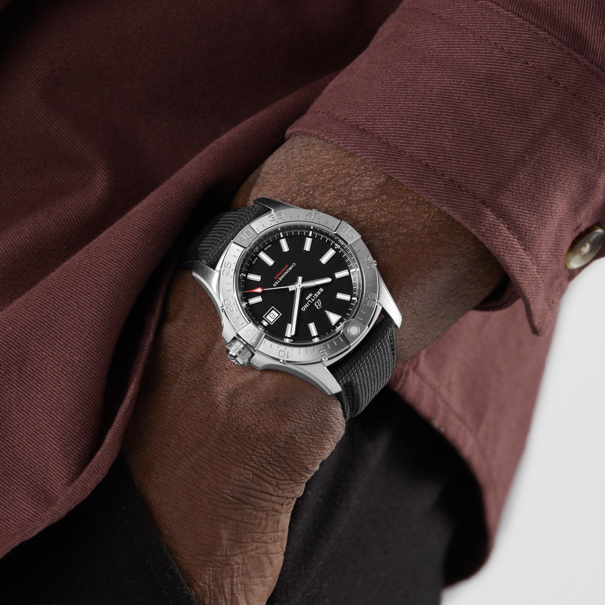 A person in a maroon shirt and black pants, hand in pocket, wears a silver and black Breitling Avenger Automatic 42 wristwatch with a stainless steel case and black strap.