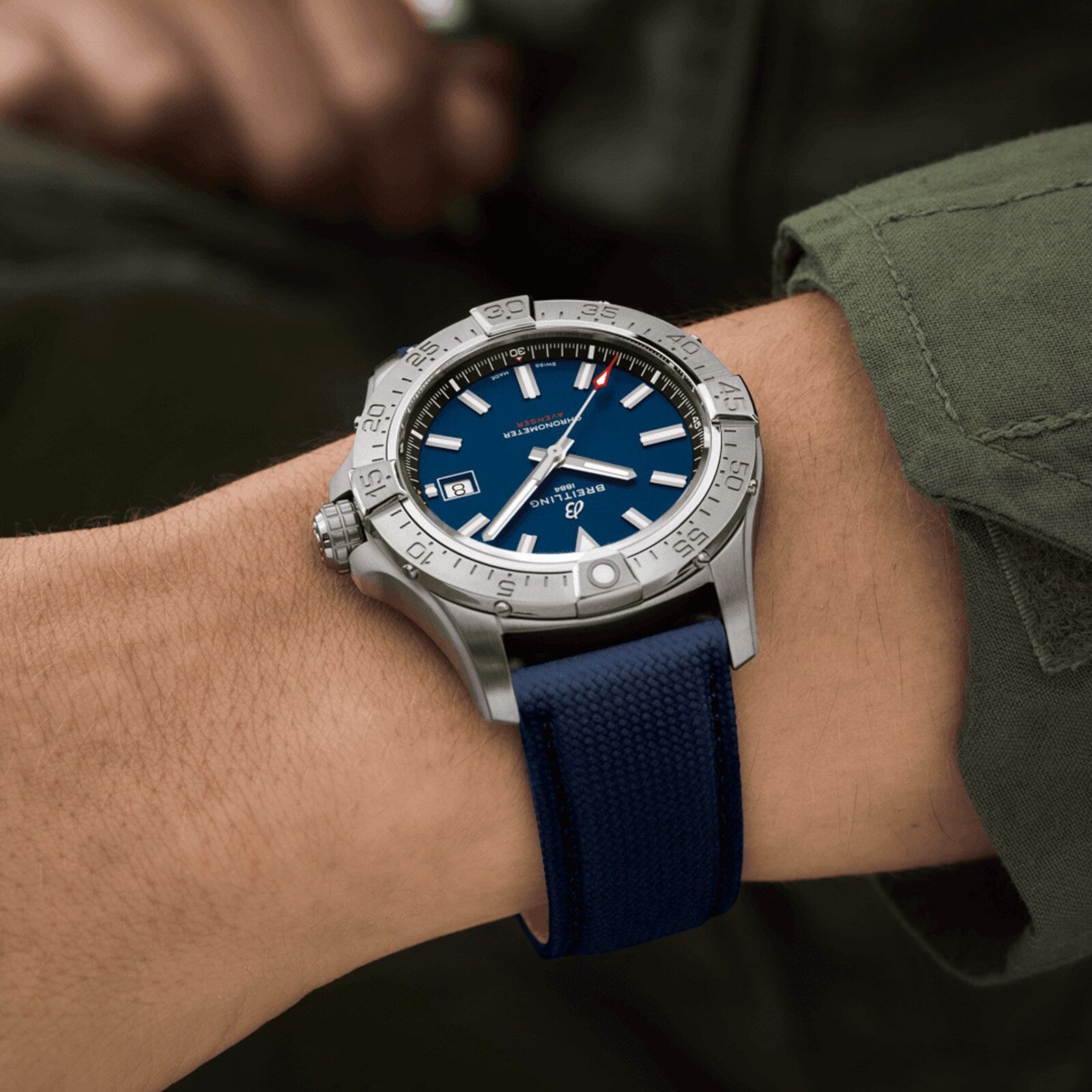 A person wearing a Breitling Avenger Automatic 42 with a blue dial and strap, and a 42mm stainless steel case, shows the time at 10:08 and date, standing out against an olive green sleeve.