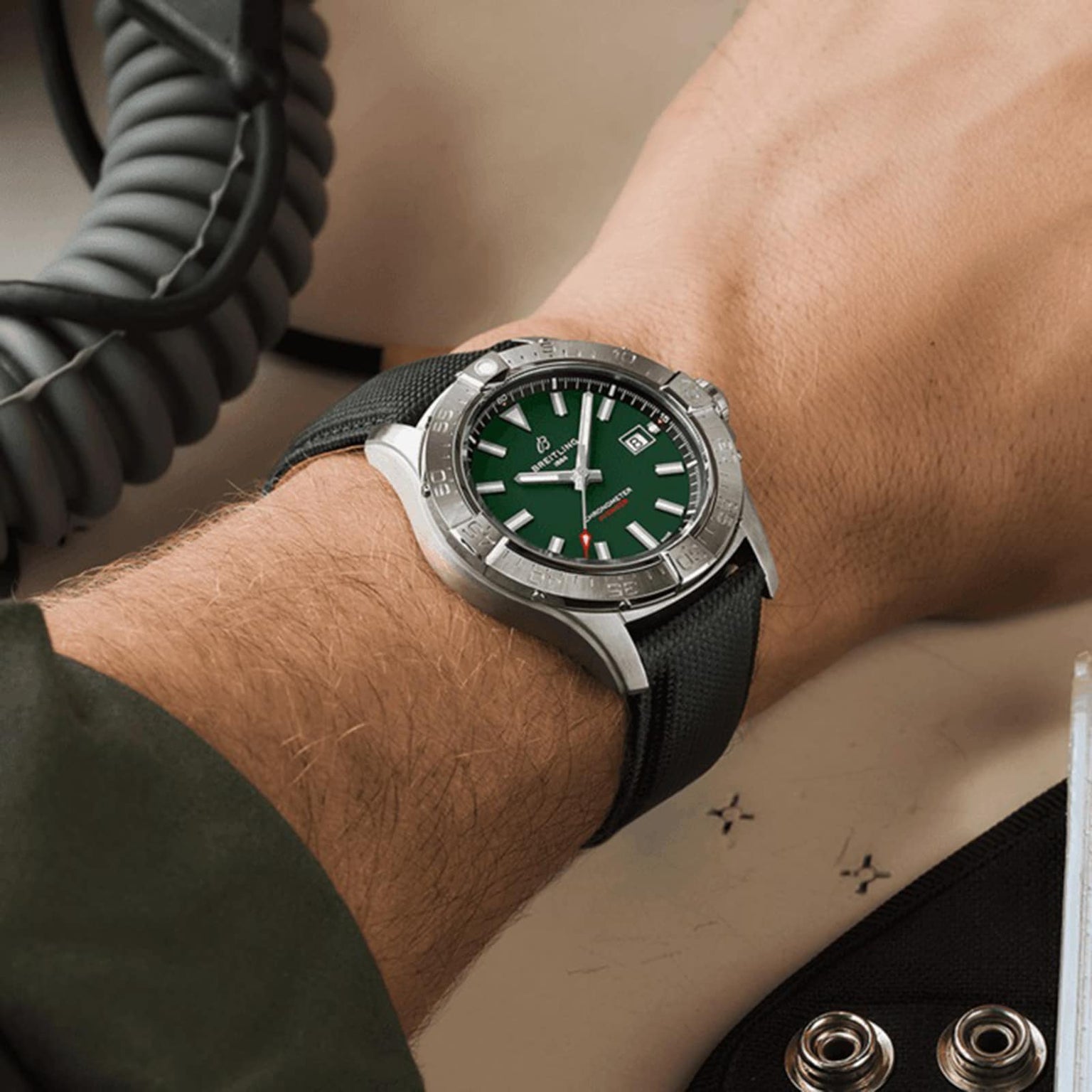 A person wears a Breitling Avenger Automatic 42 with a silver case, green face, and black military leather strap, showing date and time amid technical equipment.