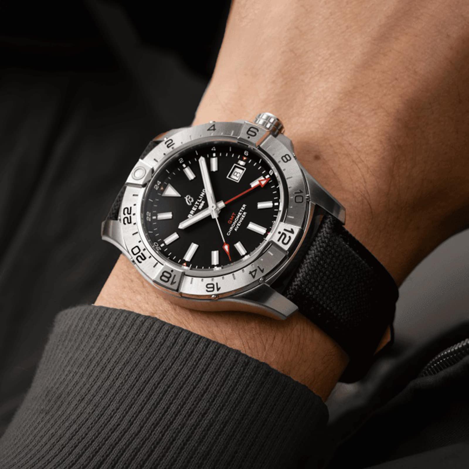 A persons wrist wearing a Breitling Avenger Automatic GMT 44 with stainless steel case, black dial, white and red hands, date display, and a military-grade leather strap.