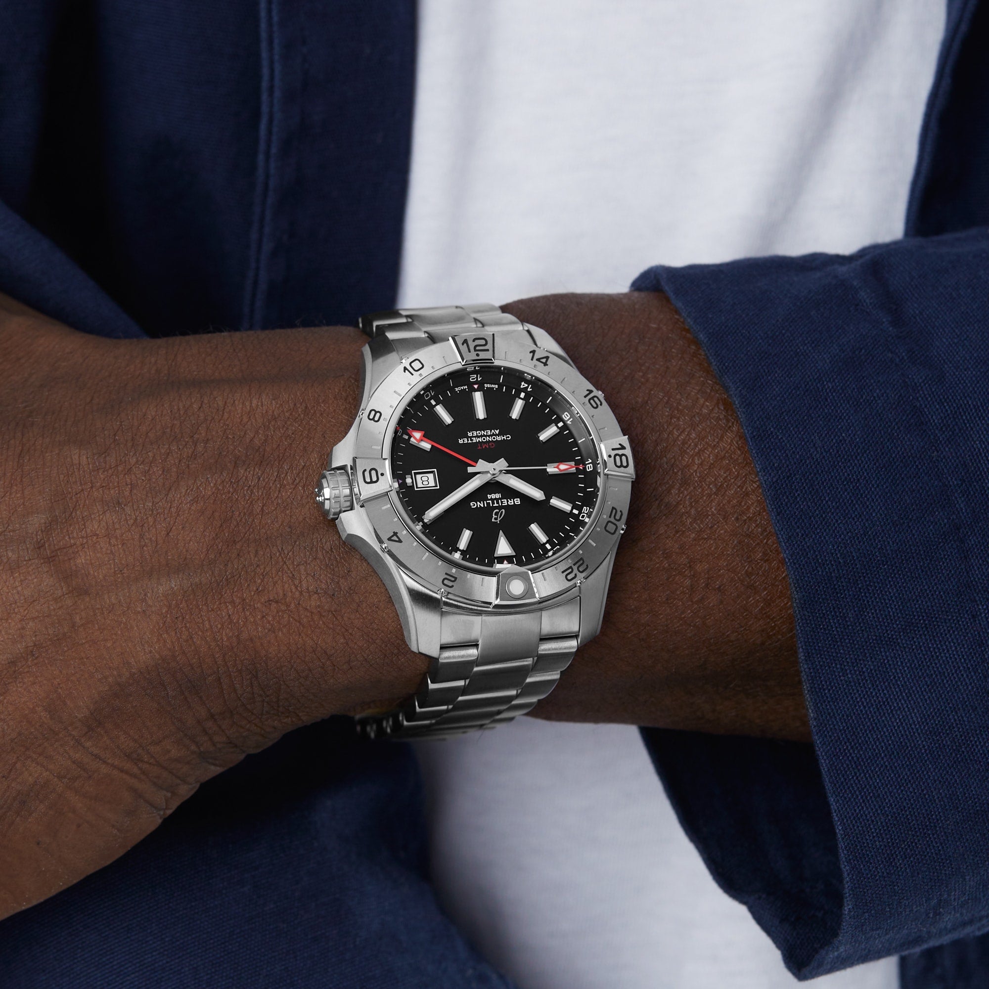 A person wearing a Breitling Avenger Automatic GMT 44 wristwatch in silver stainless steel with a black dial and date display, paired with a navy jacket and white shirt.