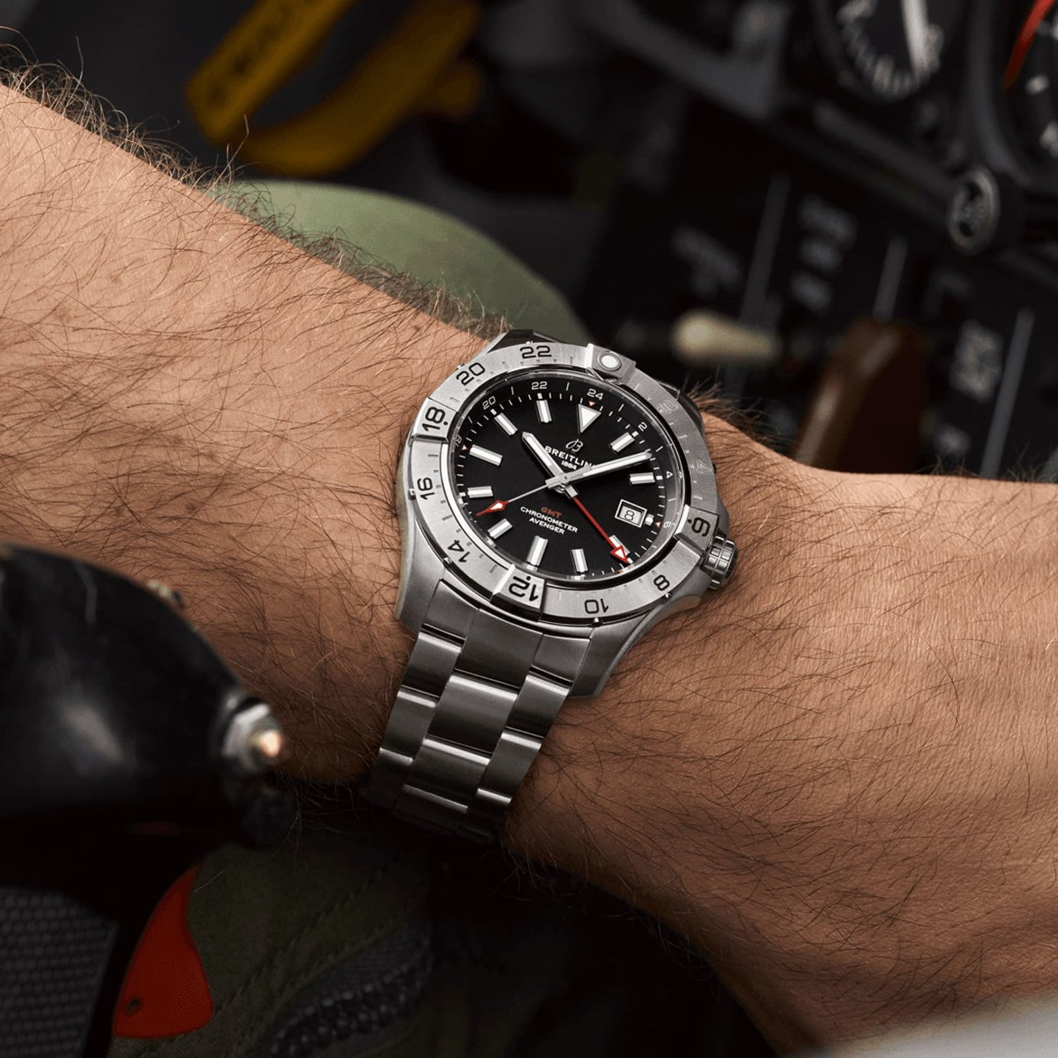 A Breitling Avenger Automatic GMT 44 wristwatch with a black dial, date display, and rotating bezel is worn on a left wrist, with aircraft controls in the background.