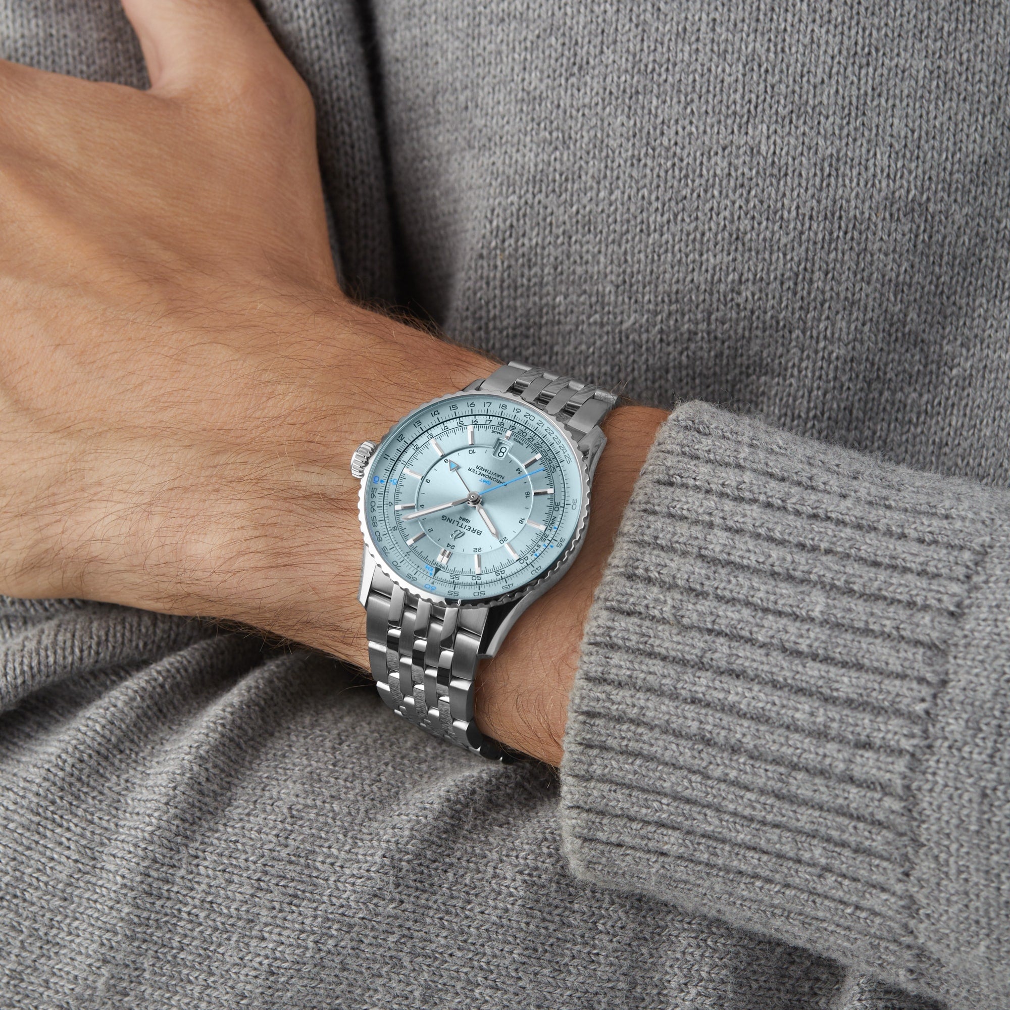 A person in a gray knit sweater wears a Breitling Navitimer Automatic GMT 41 wristwatch with a silver-tone case, light blue dial, and metal band on their left wrist.
