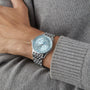 A person in a gray knit sweater wears a Breitling Navitimer Automatic GMT 41 wristwatch with a silver-tone case, light blue dial, and metal band on their left wrist.