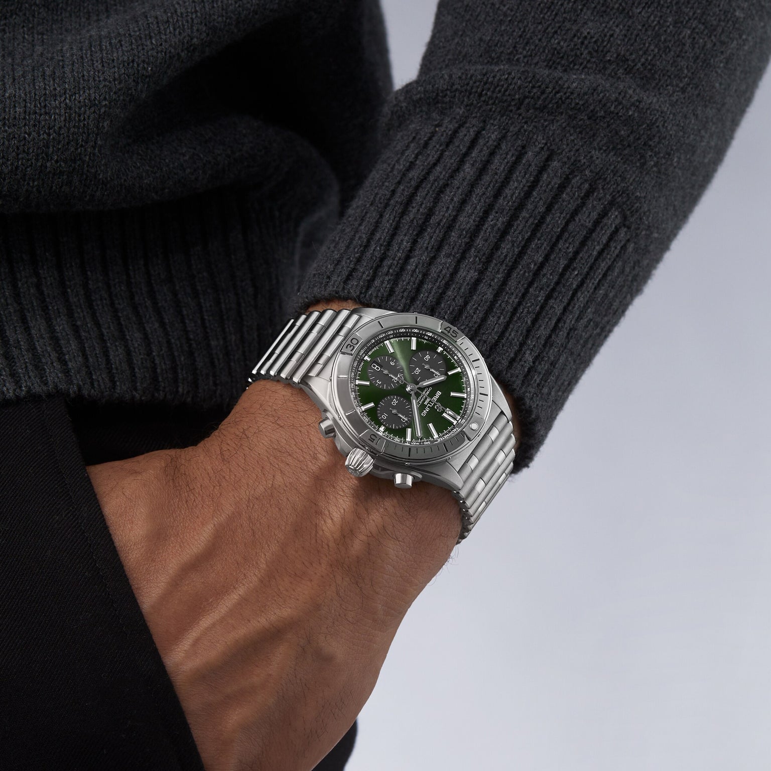 A person wearing the Breitling Chronomat B01 42 The Watches Of Switzerland Group Centenary Exclusive, a limited edition with silver case and green dial featuring chronograph functions, dressed in a dark sweater with their hand in their pocket.