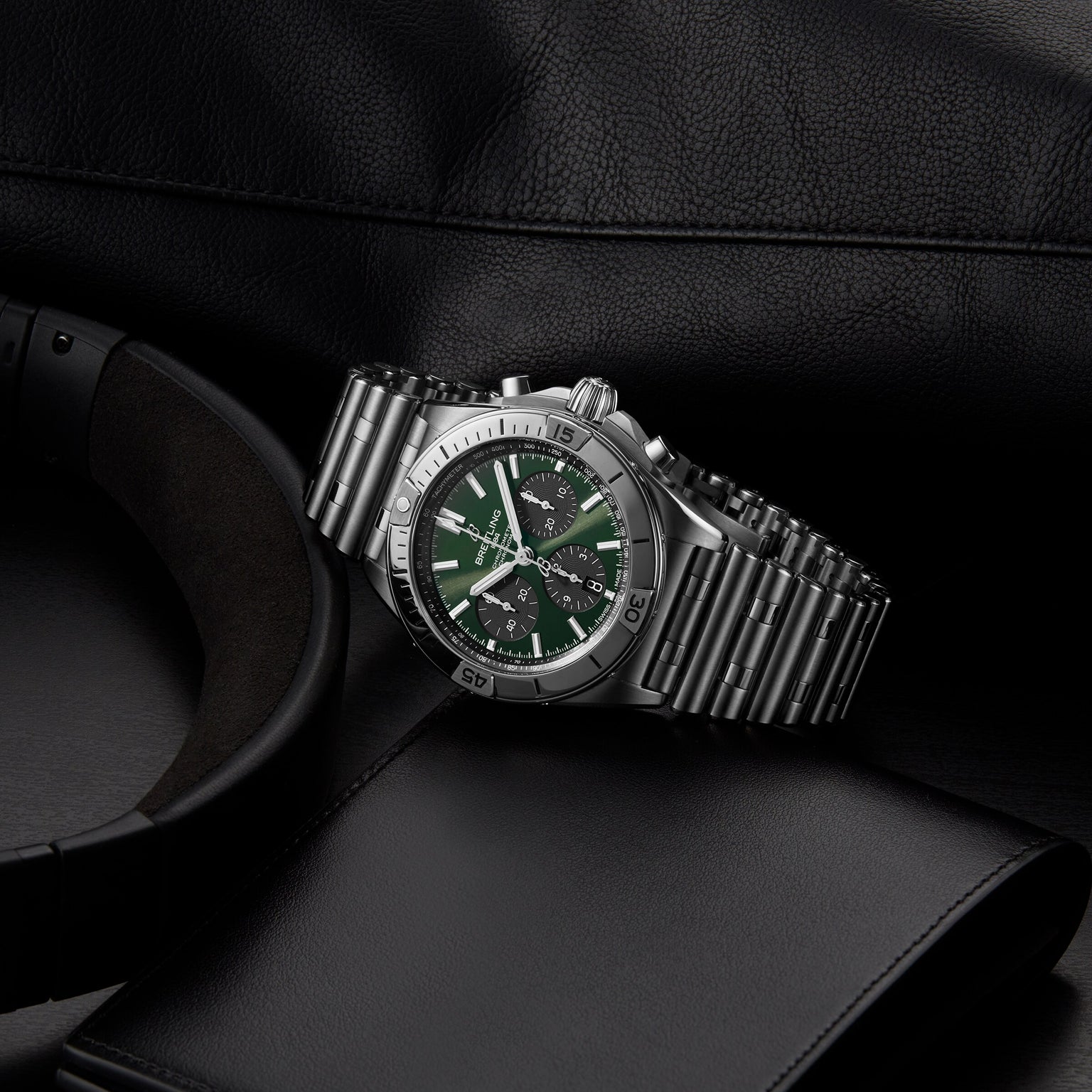 A Breitling Chronomat B01 42 The Watches Of Switzerland Group Centenary Exclusive wristwatch with a green and black dial rests on a dark surface beside a black wallet, headphones, and leather item. This limited edition features the famed B01 Manufacture Calibre.