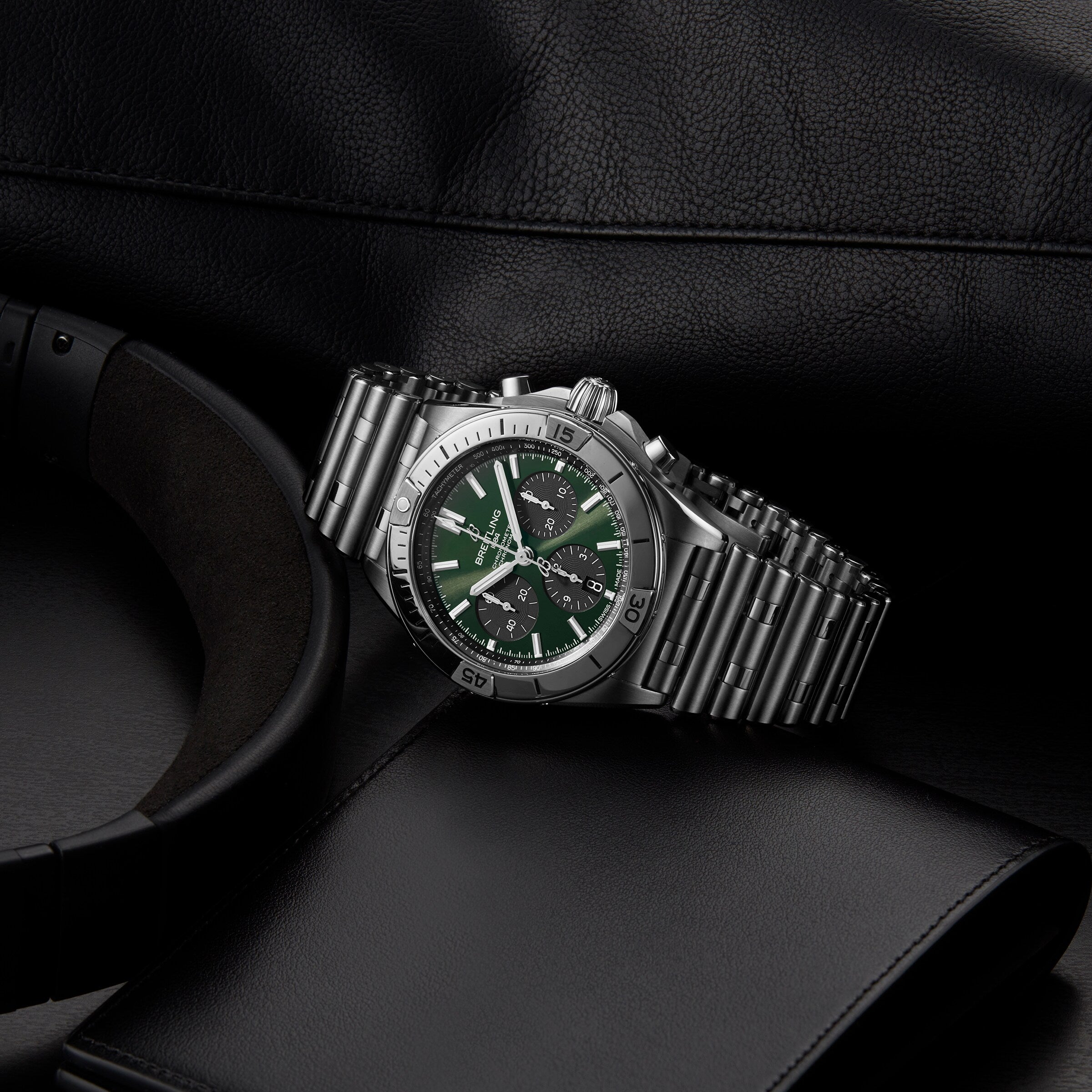 A Breitling Chronomat B01 42 The Watches Of Switzerland Group Centenary Exclusive wristwatch with a green and black dial rests on a dark surface beside a black wallet, headphones, and leather item. This limited edition features the famed B01 Manufacture Calibre.
