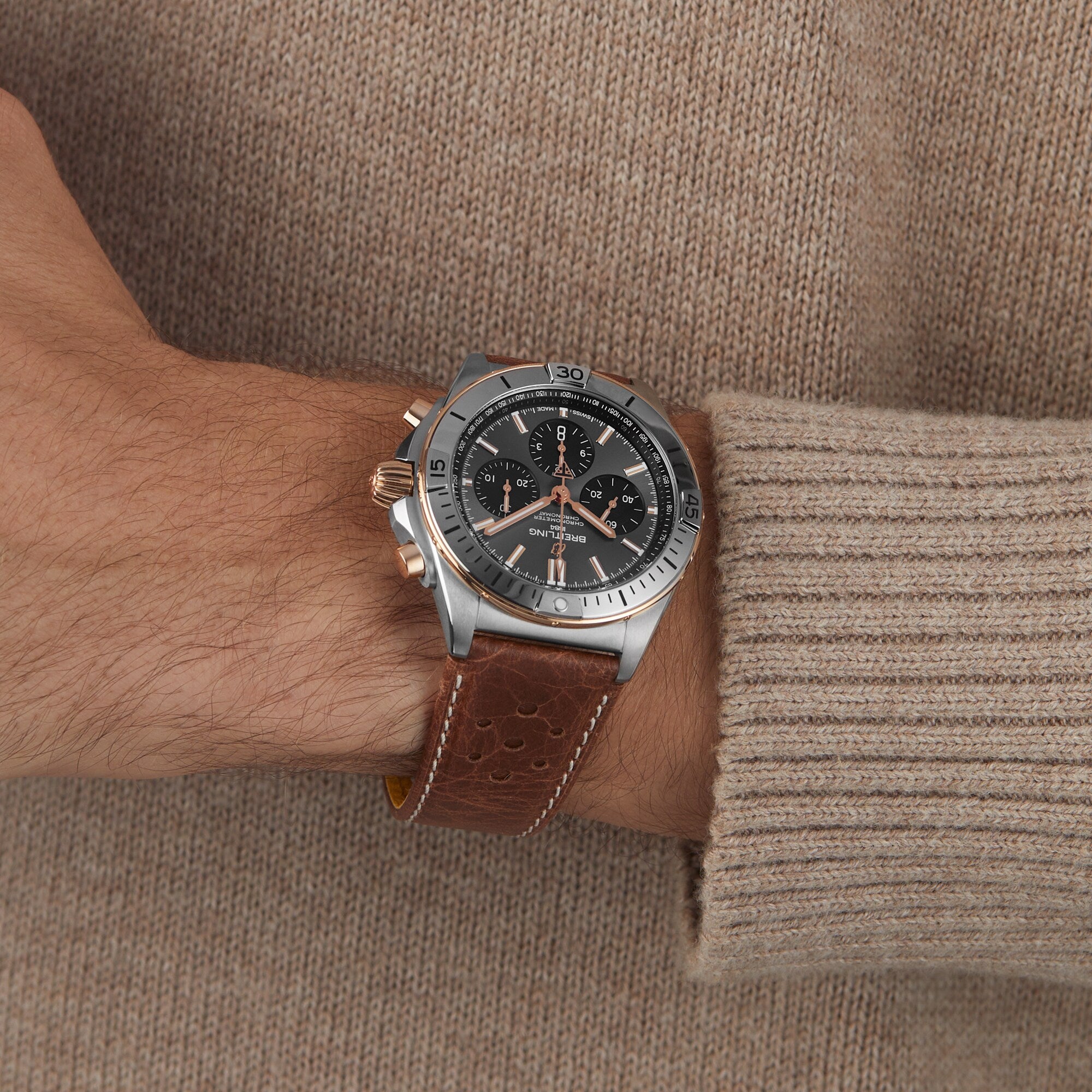 A wrist showcasing the Breitling Chronomat B01 42 Triumph, featuring a brown leather strap, black dial, and silver bezel, worn with a beige sweater.