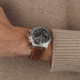 A wrist showcasing the Breitling Chronomat B01 42 Triumph, featuring a brown leather strap, black dial, and silver bezel, worn with a beige sweater.