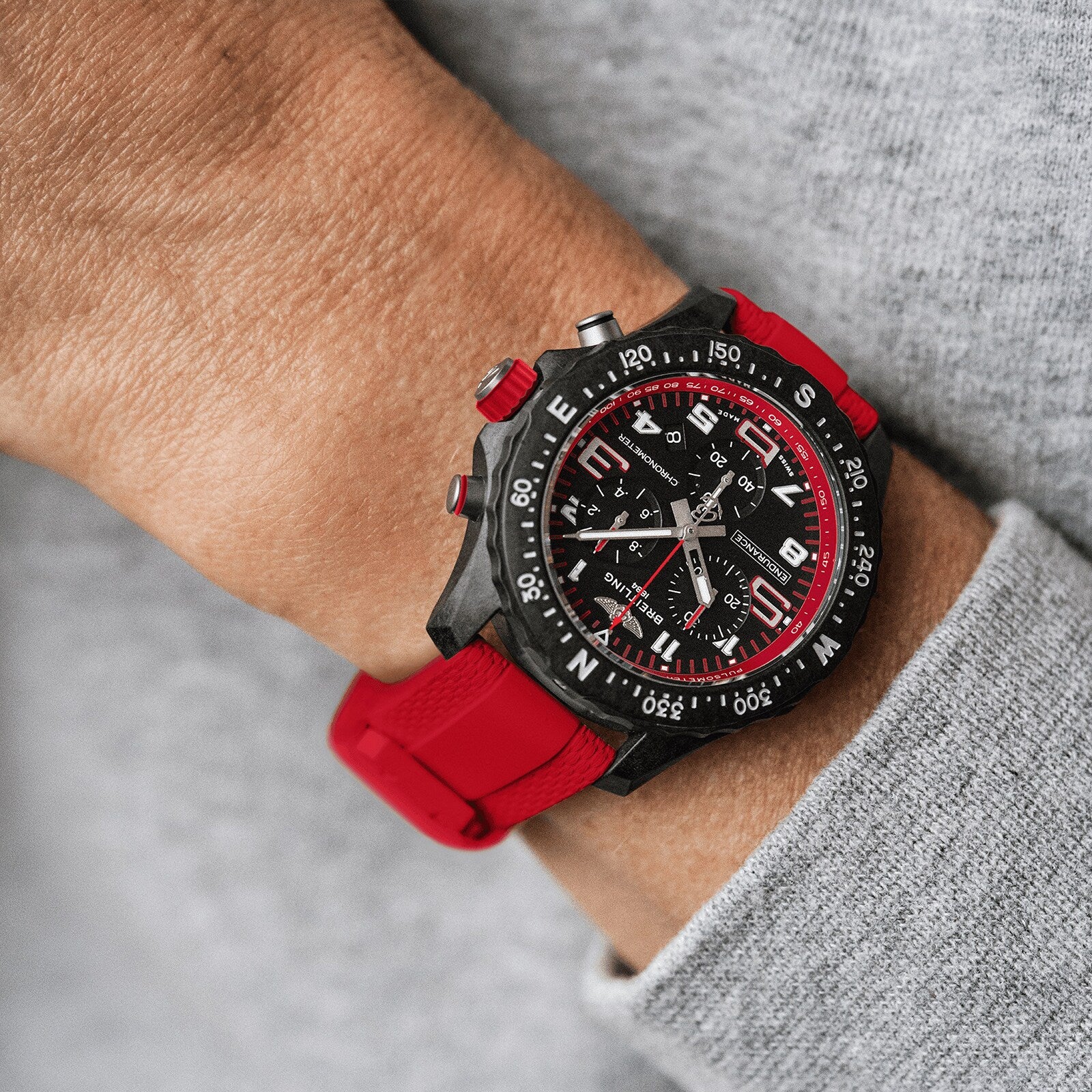 A person wearing the Breitling Endurance Pro 38, featuring a black ultra-light case, red rubber strap, and white bezel markings, shown on their wrist against a grey sleeve background.