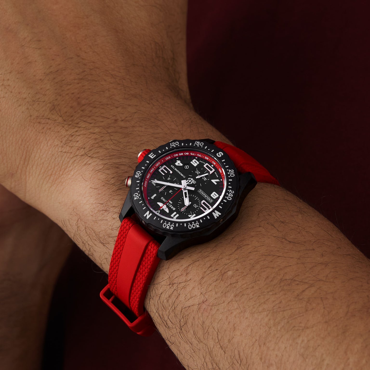 A wrist wears the Breitling Endurance Pro 38, a black and red athleisure watch with a compass bezel, red strap, and multiple dials, set against a brownish background.