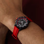A wrist wears the Breitling Endurance Pro 38, a black and red athleisure watch with a compass bezel, red strap, and multiple dials, set against a brownish background.