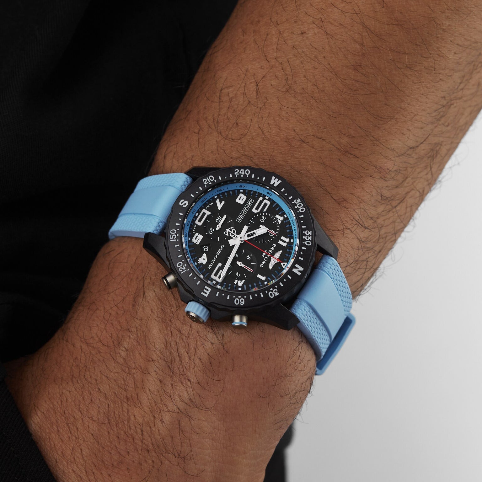 A person wears a Breitling Endurance Pro 38 watch with a black and blue analog dial, compass bezel, blue strap, and ultra-light Breitlight case on their left wrist.