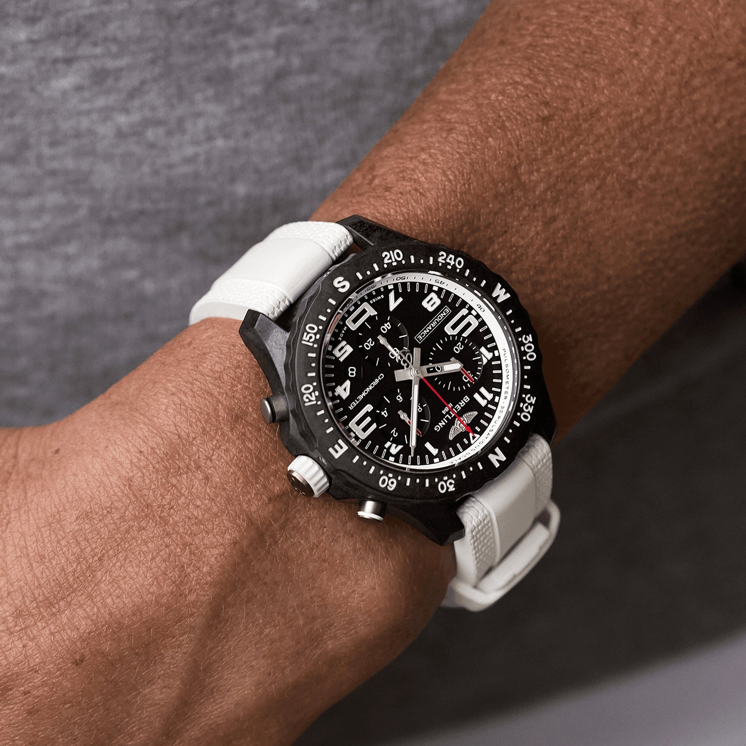 A person wears a Breitling Endurance Pro 38 athleisure watch with a white strap, Breitlight case, compass bezel, and multiple dials against a grey background.