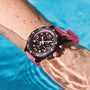 A wrist wearing a Breitling Endurance Pro 38 athleisure watch with a black and red case and pink strap is submerged in clear blue water.