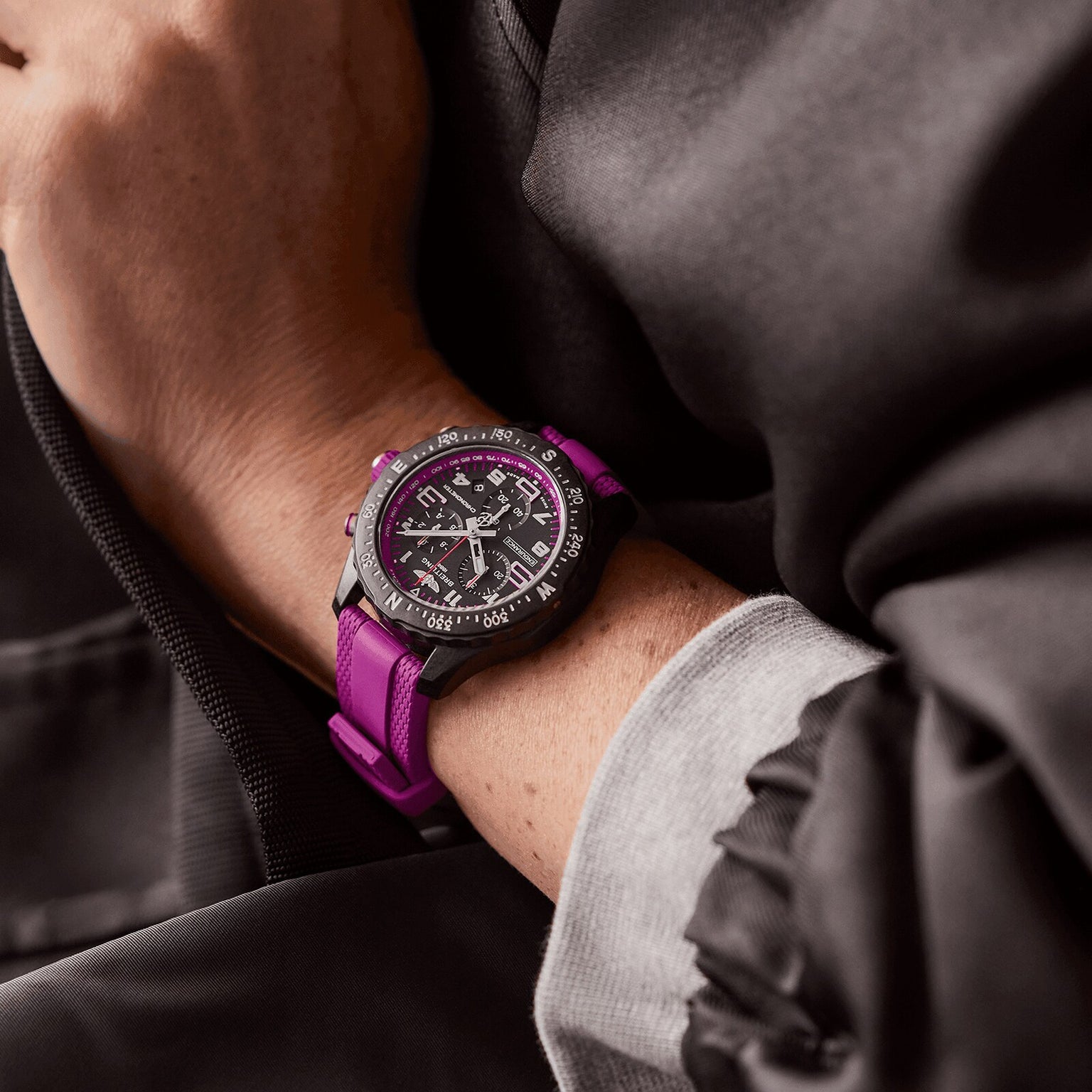 A person wears the Breitling Endurance Pro 38, a black and silver athleisure watch with a purple strap, against a dark jacket and light gray sleeve.