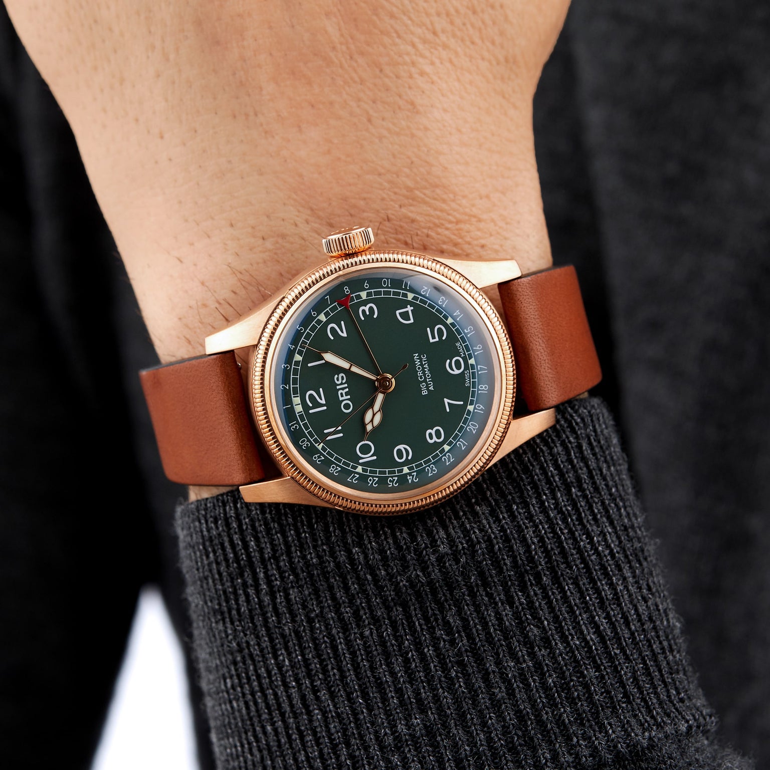 A person wears an Oris Big Crown Pointer Date 80th Anniversary Edition with a green dial, gold case, brown leather strap, and Arabic numerals over a dark long-sleeve top.