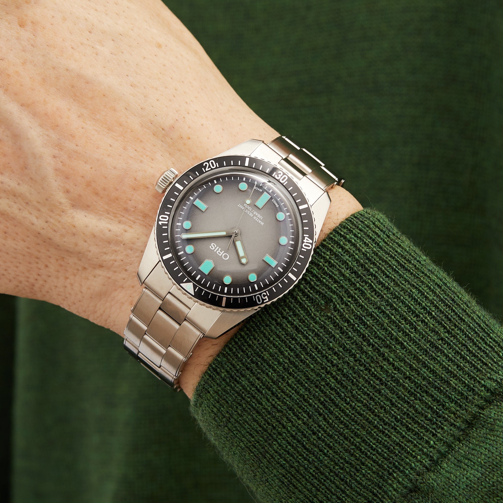 An Oris Divers Sixty‑Five silver wristwatch with a black bezel, worn on a persons wrist paired with a green sweater.