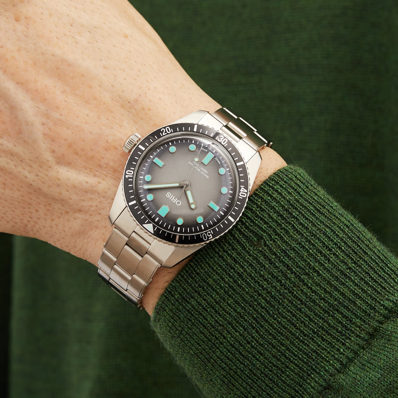 An Oris Divers Sixty‑Five silver wristwatch with a black bezel, worn on a persons wrist paired with a green sweater.