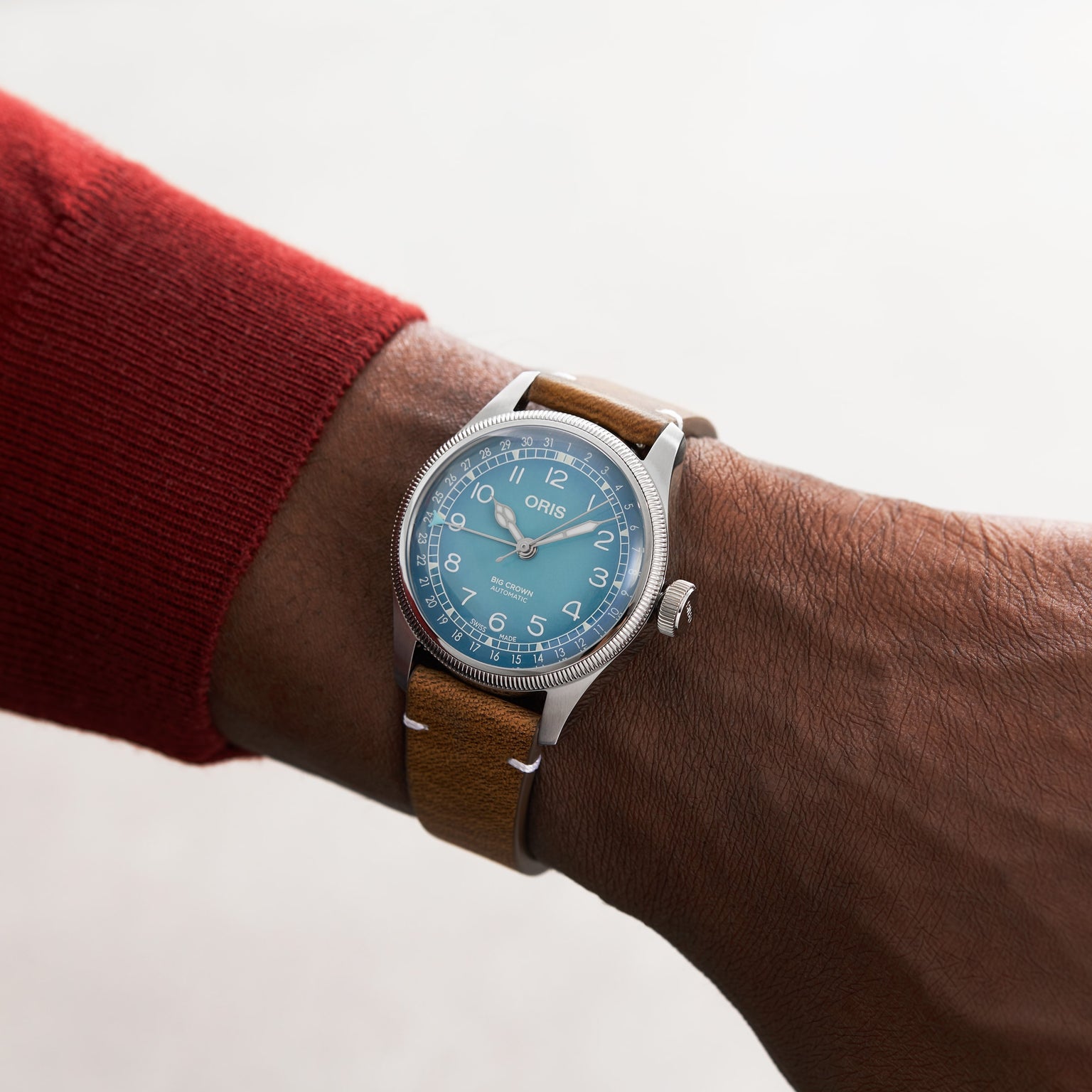 A person wearing an Oris Big Crown Oris X Cervo Volante watch with a light blue dial and brown leather strap, paired with a red long-sleeve top.