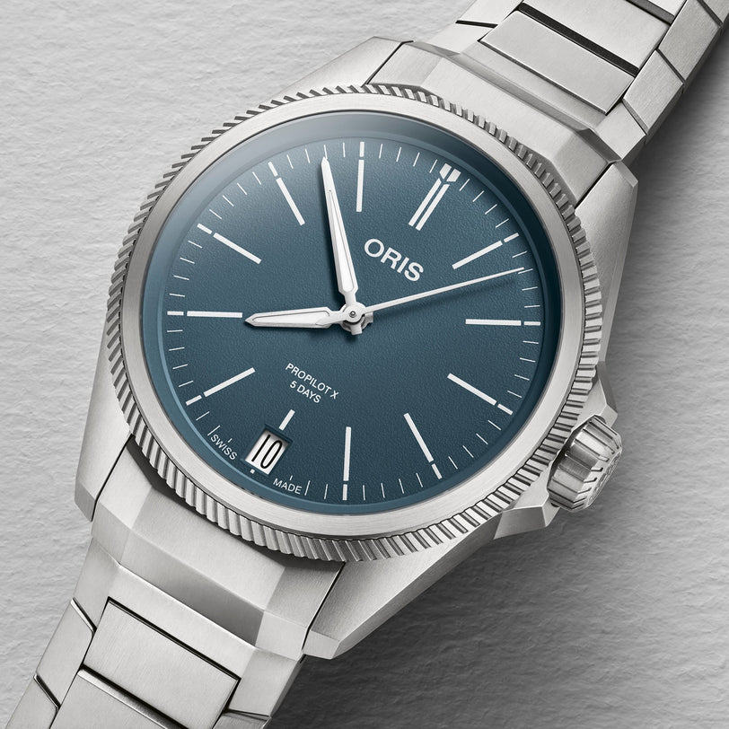 The Oris ProPilot X Calibre 400 watch features a blue dial, silver hour markers, date at 6 o’clock, and a stainless steel bracelet—reflecting aviation heritage against a light grey background.
