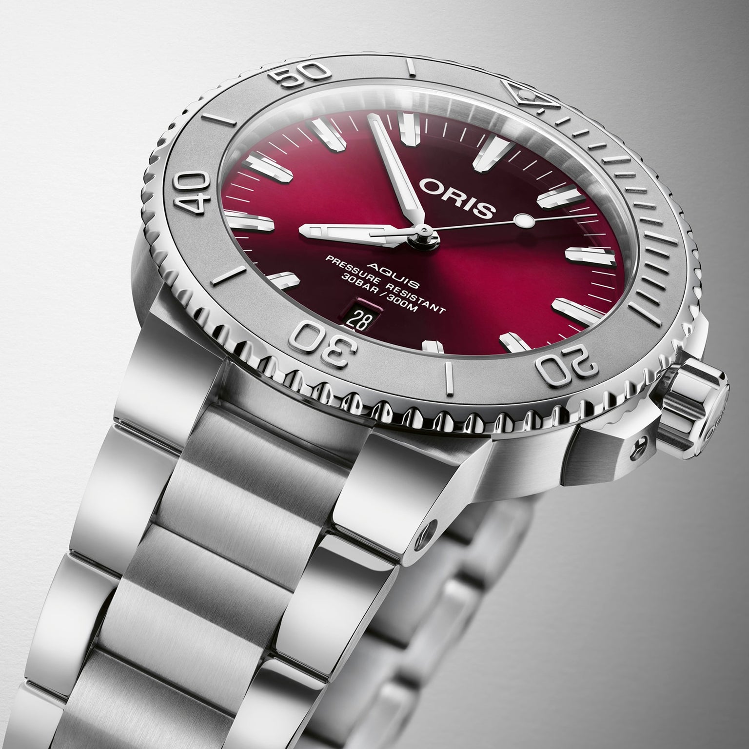 The Oris Aquis Date Relief is a stainless steel wristwatch with a red dial, silver bezel, and date display at the 6 o’clock position.