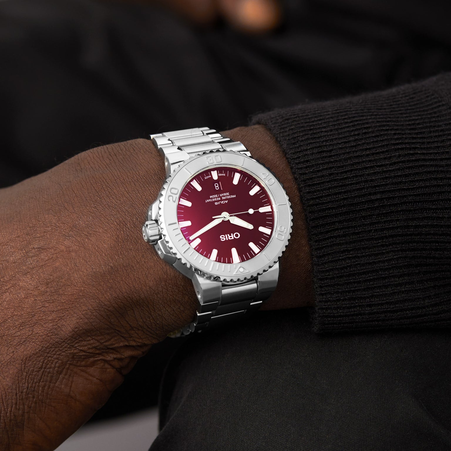 An Oris Aquis Date Relief watch with a silver case and burgundy dial, known for its precise mechanical movement, is worn on the wrist of a person dressed in a black long-sleeve shirt.