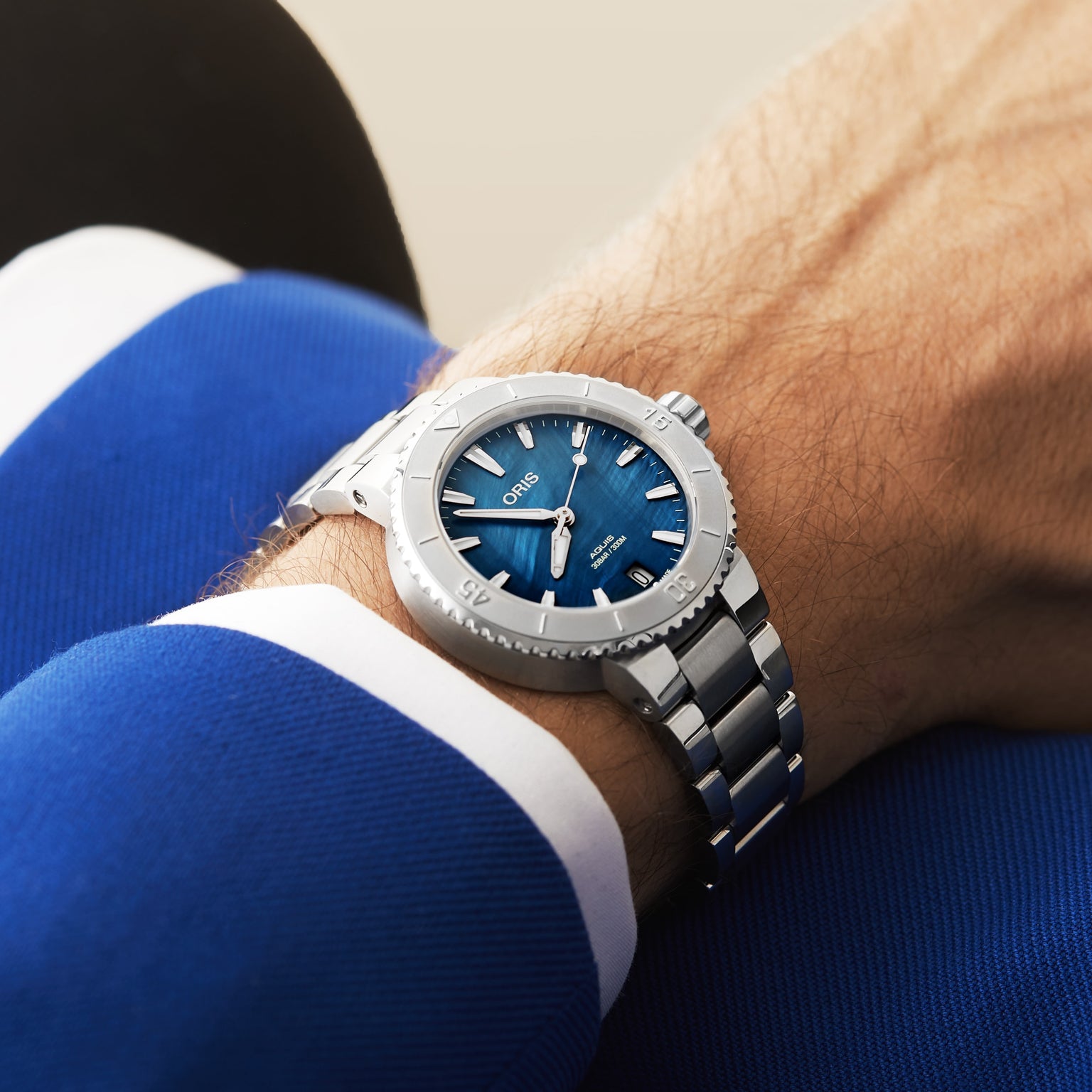 A person wearing an Oris Aquis Date watch with a blue dial and silver metal band, dressed in a blue jacket with white trim.