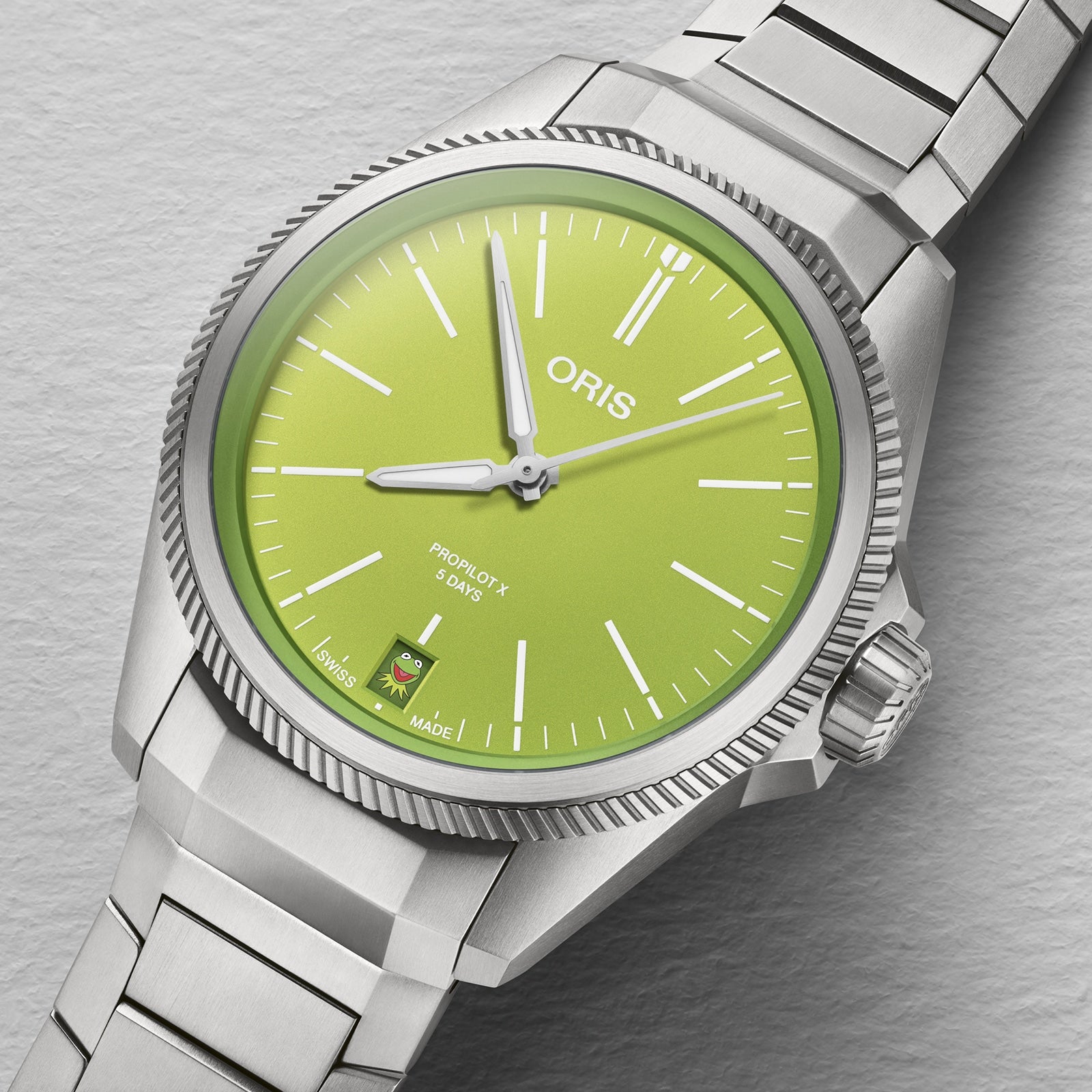 The Oris ProPilot X Kermit Edition wristwatch features a green dial, silver markers and hands, a date window at 6 o’clock, and is displayed on a textured gray surface.