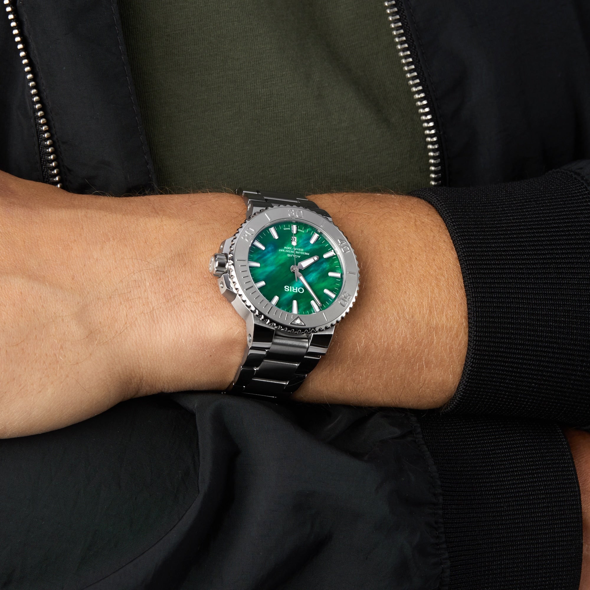 A person in a black jacket and olive green shirt crosses their arms, showing an Oris Aquis Oris X Bracenet wristwatch with a vivid green face and metal band on their left wrist.
