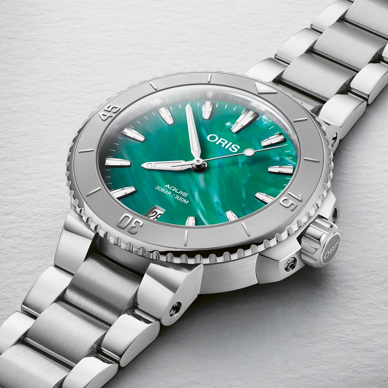 Close-up of the Oris Aquis Oris X Bracenet watch with a green mother-of-pearl dial, Super-LumiNova silver hour markers, and a stainless steel bracelet on a light textured surface.