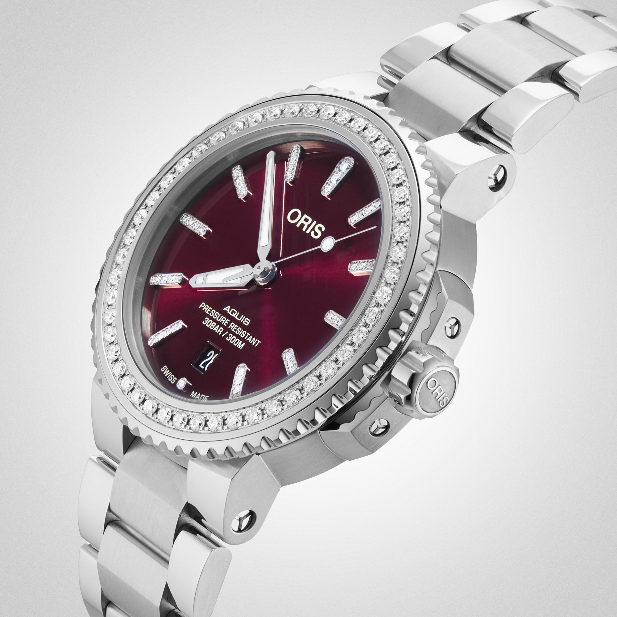 A close-up of the Oris Aquis Date Diamonds wristwatch featuring a red dial, lab-grown diamond hour markers, and a stainless steel band set against a light background.