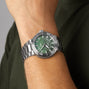 An Oris Dat Watt Limited Edition II silver wristwatch with a green dial is worn on a person’s wrist, paired with a dark green shirt.