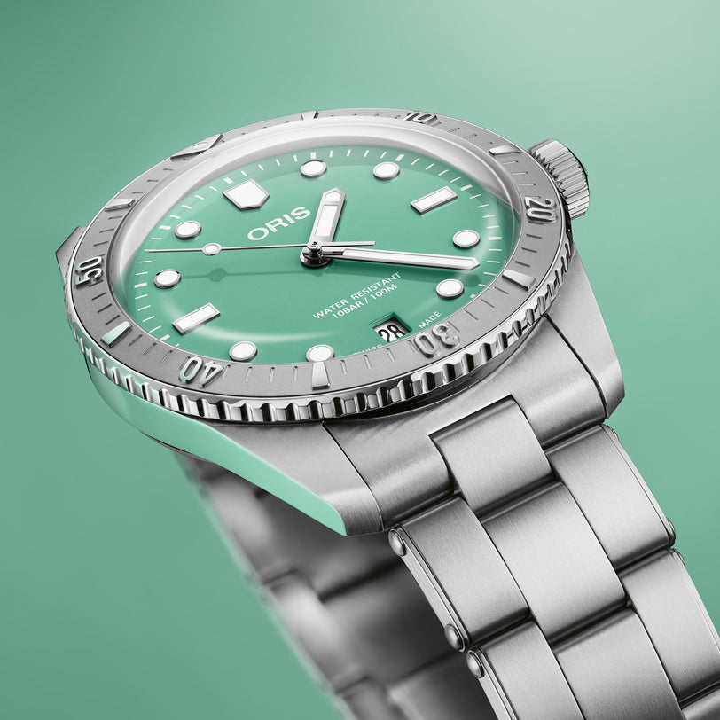 The Oris Divers Sixty‑Five Cotton Candy, a silver wristwatch with a green dial and metal band, is displayed against a matching green background.