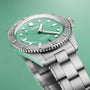The Oris Divers Sixty‑Five Cotton Candy, a silver wristwatch with a green dial and metal band, is displayed against a matching green background.