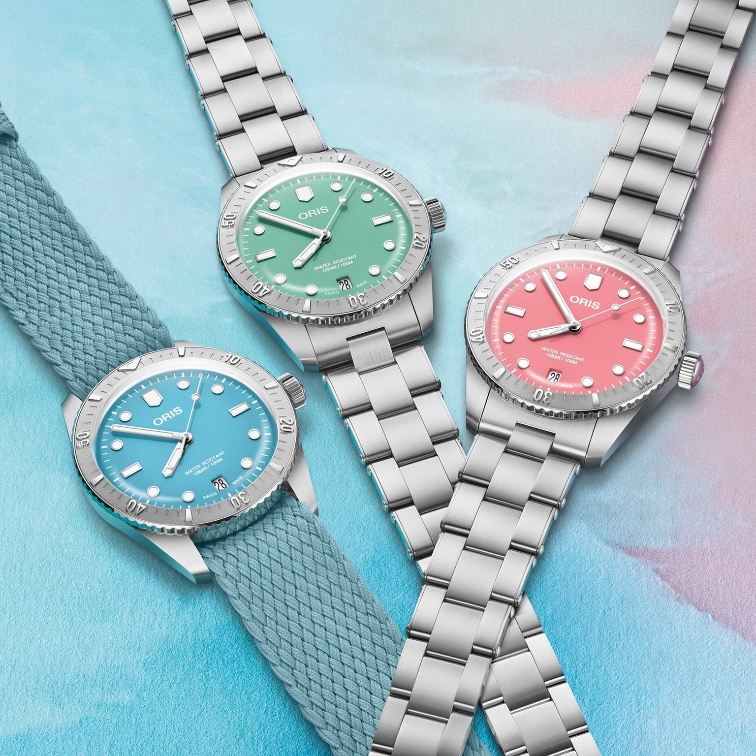 Three Oris Divers Sixty‑Five Cotton Candy watches with blue, green, and pink dials are displayed on a pastel-colored background.
