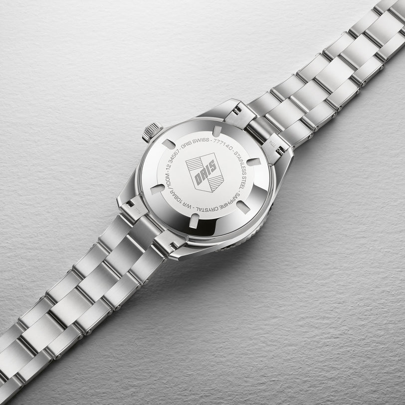 The Oris Divers Sixty-Five Cotton Candy, a silver metal wristwatch with an engraved Oris logo on the back case, rests on a textured surface.