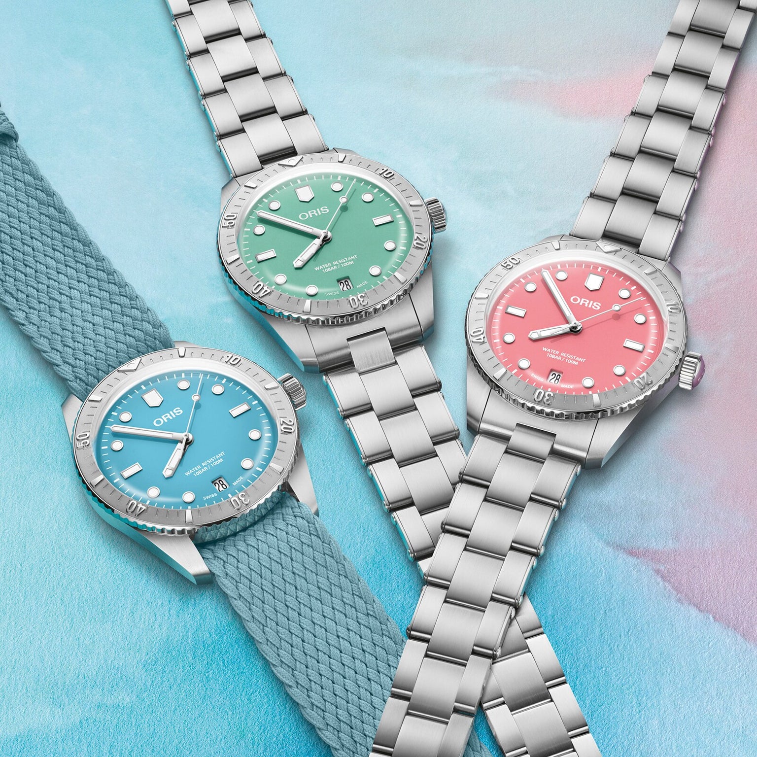 Three Oris Divers Sixty‑Five Cotton Candy watches—with blue, green, and pink dials—are displayed on a pastel background; two feature metal straps, while one has a fabric strap.