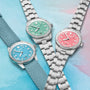 Three Oris Divers Sixty‑Five Cotton Candy watches—with blue, green, and pink dials—are displayed on a pastel background; two feature metal straps, while one has a fabric strap.