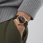 A person wearing the Oris Divers Sixty‑Five Date gold watch with a black strap and a grey sweater, hand in olive green pants pocket.