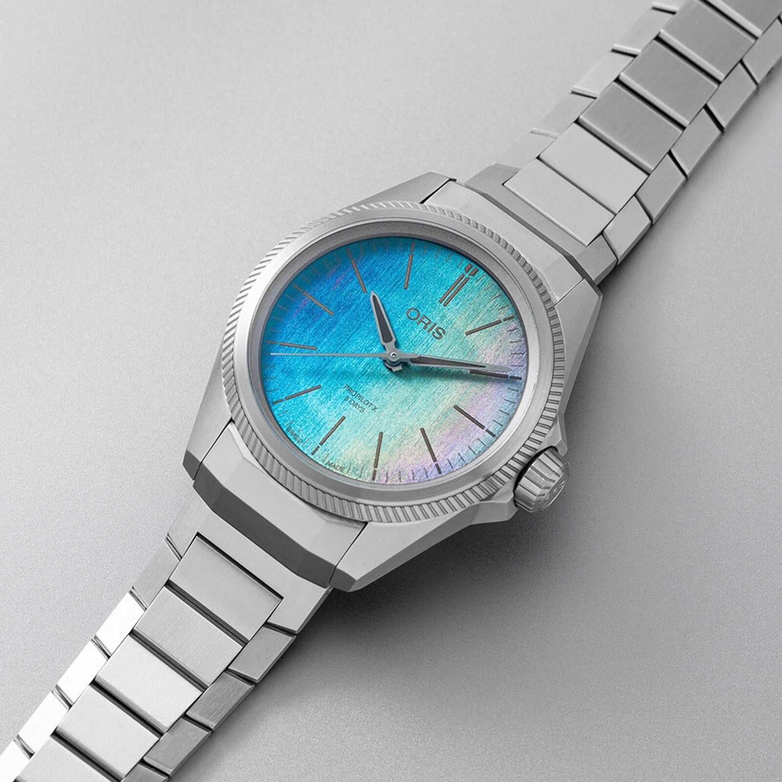 The Oris ProPilot X Calibre 400 Laser is a stainless steel wristwatch featuring a blue and purple gradient dial created with advanced laser technology, silver hour markers, and a metal link band, displayed on a light gray surface.