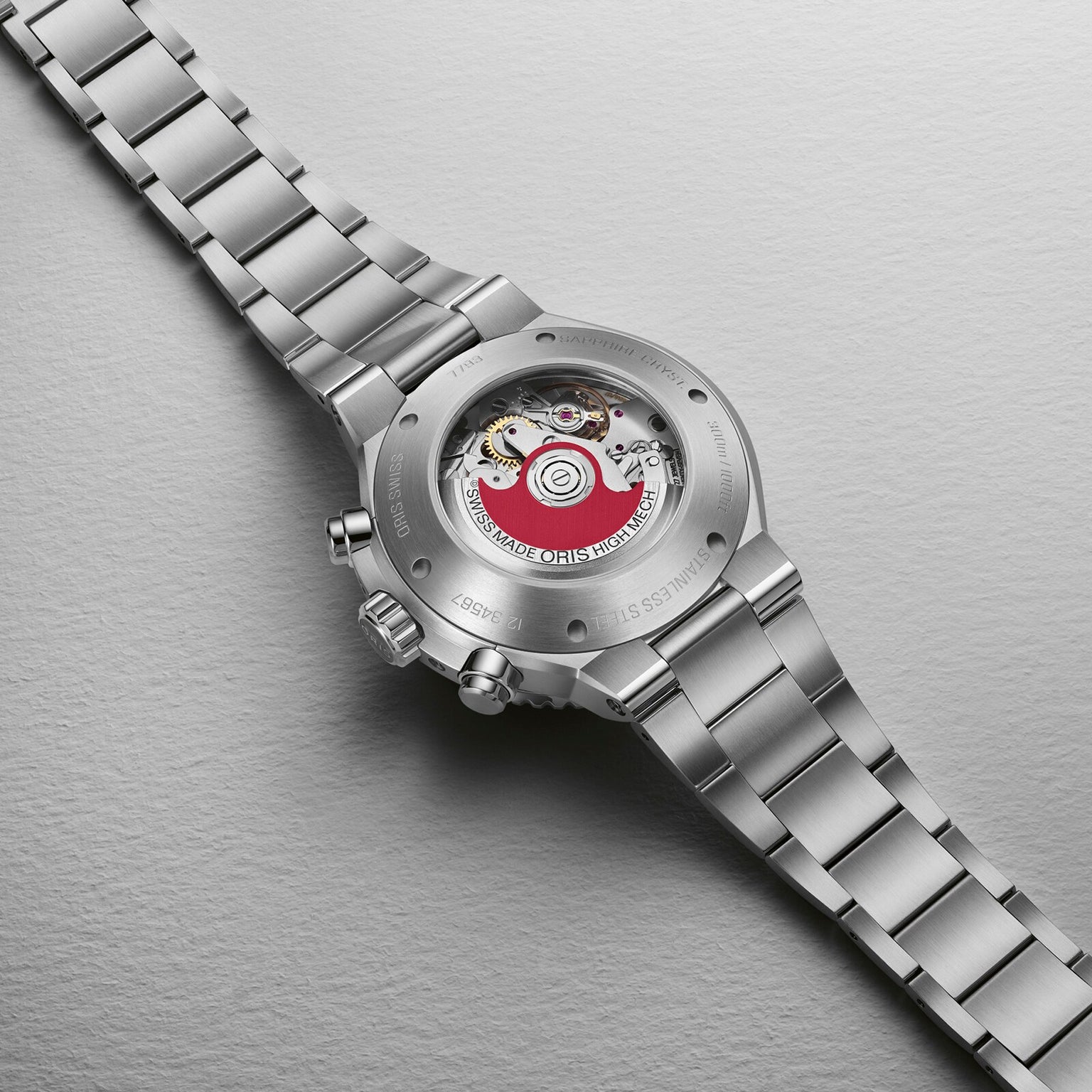 The Oris Aquis Chronograph features a stainless steel case, ceramic bezel, and a visible caseback displaying its mechanical movement and signature red rotor, shown laid flat on a textured light surface.