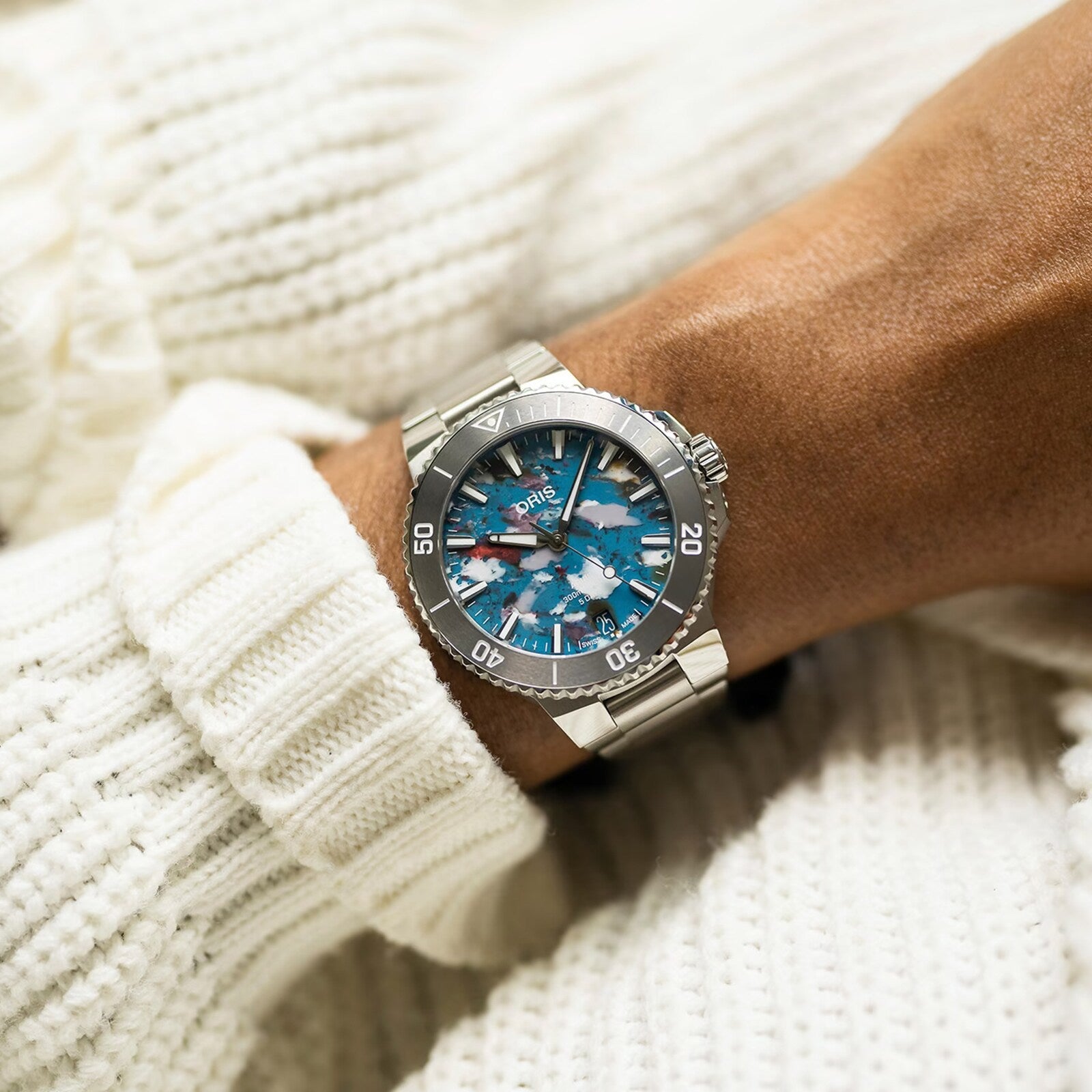A close-up of a person wearing an Oris Aquis Date Upcycle Calibre 400, featuring a silver case and blue-pink patterned dial, styled over a white knit sweater cuff—highlighting refined taste in mechanical watches.