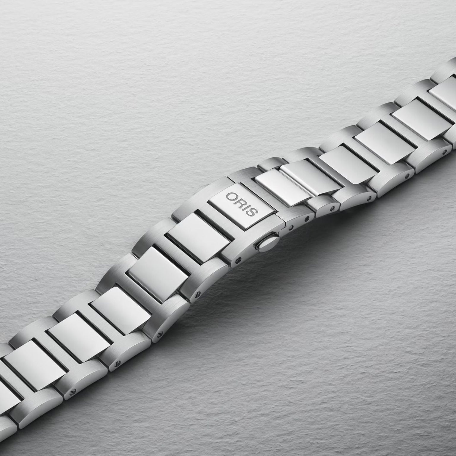 A close-up of the Oris Big Crown Pointer Date Calibre 403 stainless steel bracelet and clasp, set against a textured light grey background, showcasing its automatic movement and signature design elements.
