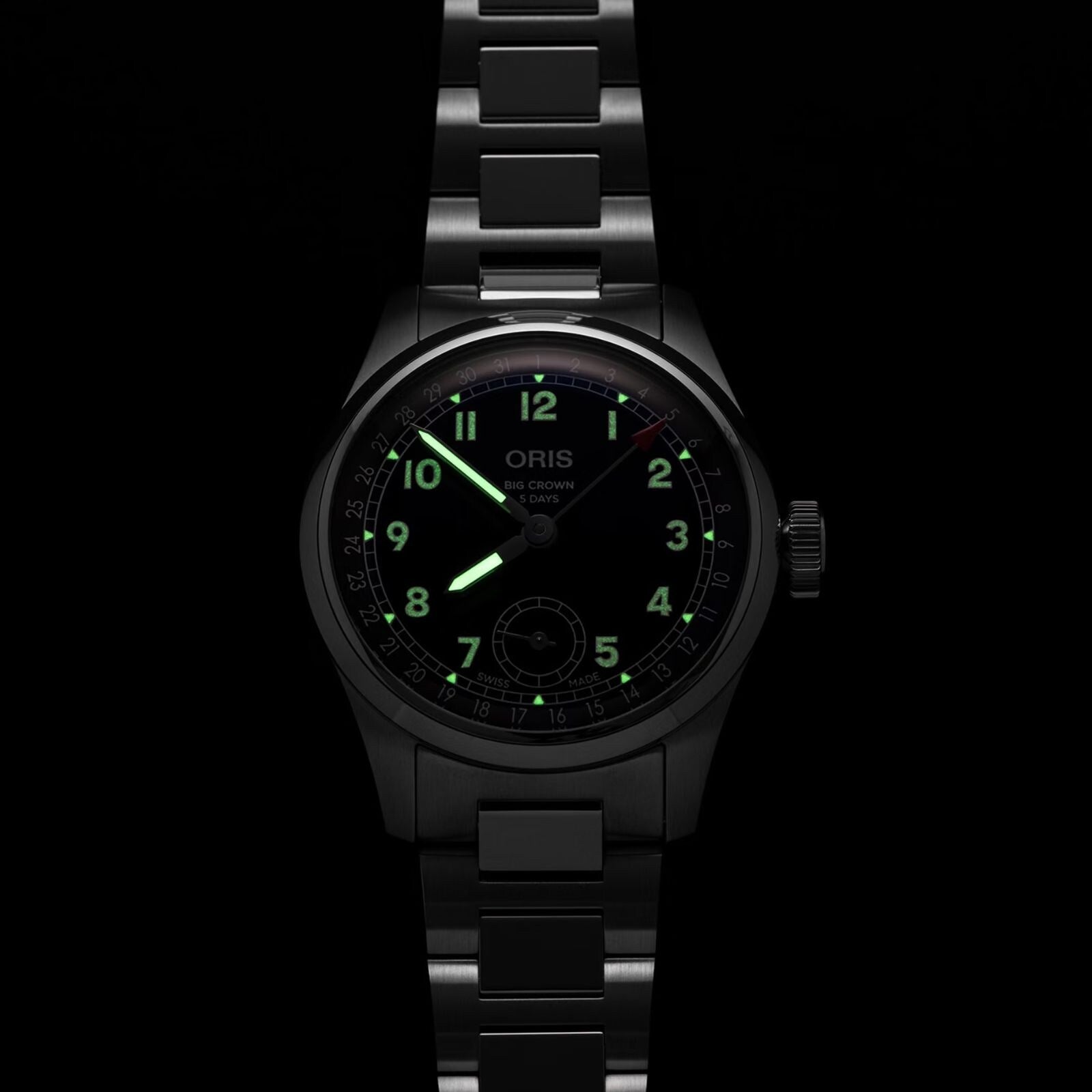 A black metal Oris Big Crown Pointer Date Calibre 403 wristwatch with luminous green numerals and hands is displayed in a dark setting, its midnight blue dial highlighting the brand’s signature automatic movement.