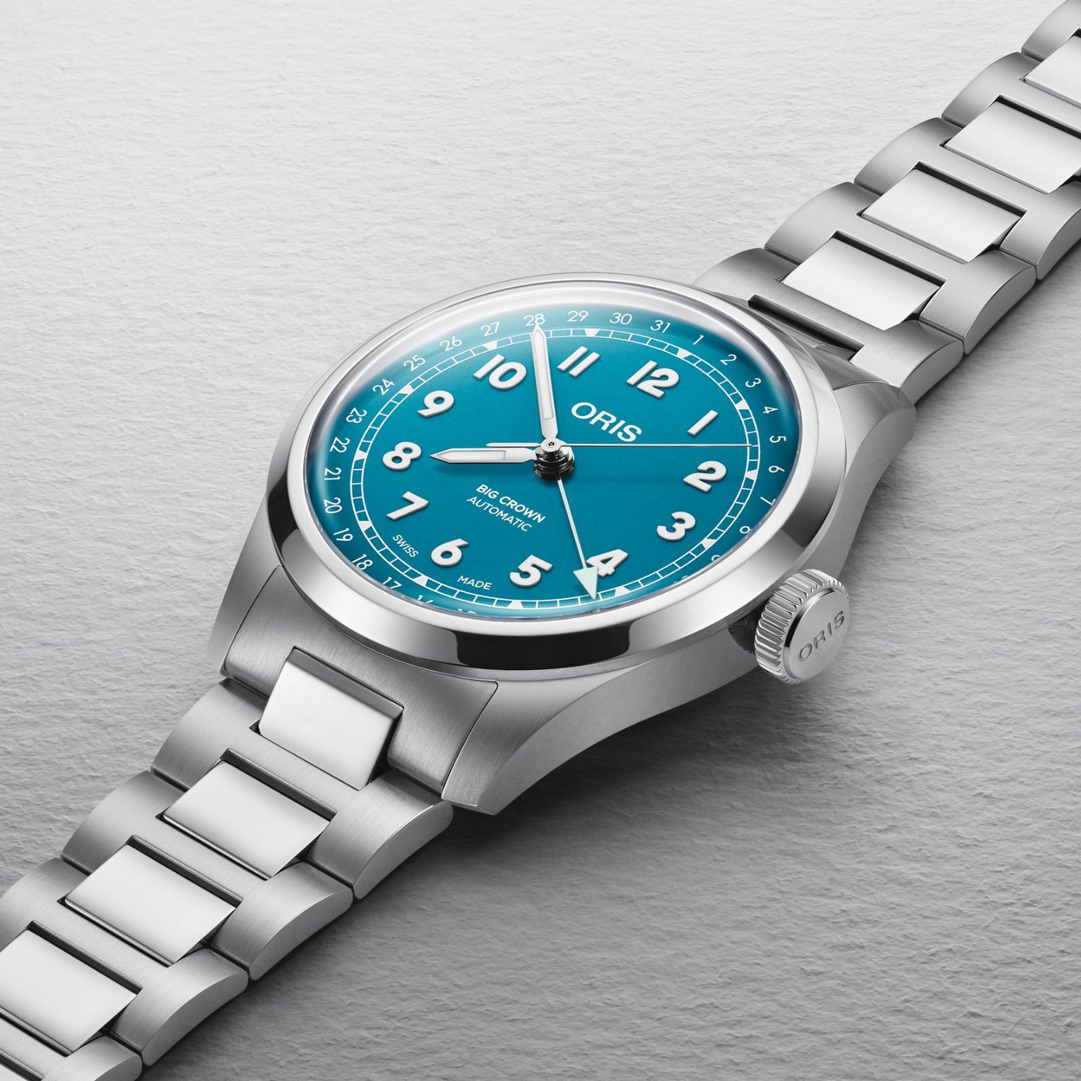 The Oris Big Crown Pointer Date wristwatch, featuring a turquoise blue dial with white numerals and a metal link bracelet, is displayed on a light gray textured surface.