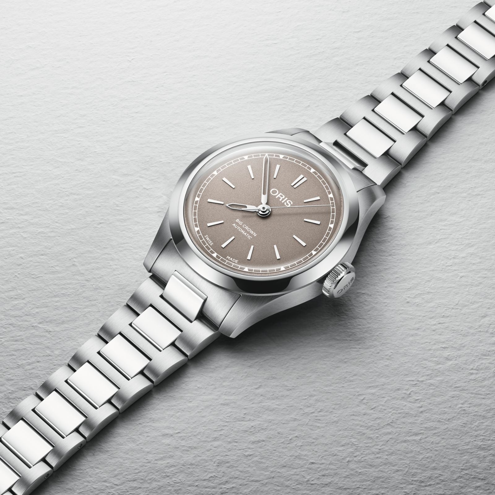The Oris Big Crown wristwatch features a silver metal case, taupe dial, and matching metal bracelet, displayed on a light gray surface.